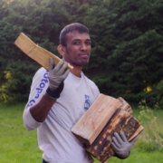 A man holding firewood in his arms while standing outdoors. He is wearing a long-sleeved shirt and gloves. The background features a green, lush garden with trees and grass.