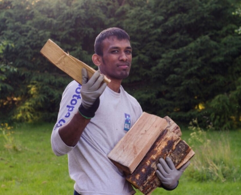 a-man-holding-firewood-in-his-arms-while-standing-outdoors-he-is-wearing-a-long-sleeved-shirt-and-gloves-the-background-features-a-green-lush-garden-with-trees-and-grass-stockpack-unsplash.jpg A man holding firewood in his arms while standing outdoors. He is wearing a long-sleeved shirt and gloves. The background features a green, lush garden with trees and grass.